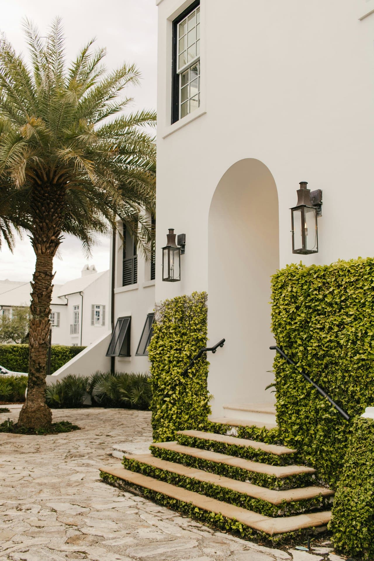 A white-stucco entry at Alys Beach — arched doorway, gas lanterns, and clipped hedges beside stone steps.