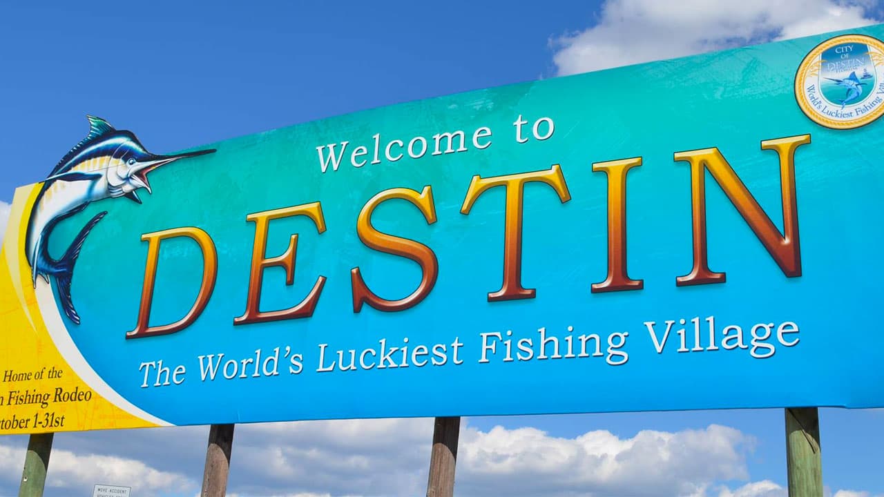 A roadside welcome sign reading 'Welcome to DESTIN — The World's Luckiest Fishing Village' against a blue Emerald Coast sky.