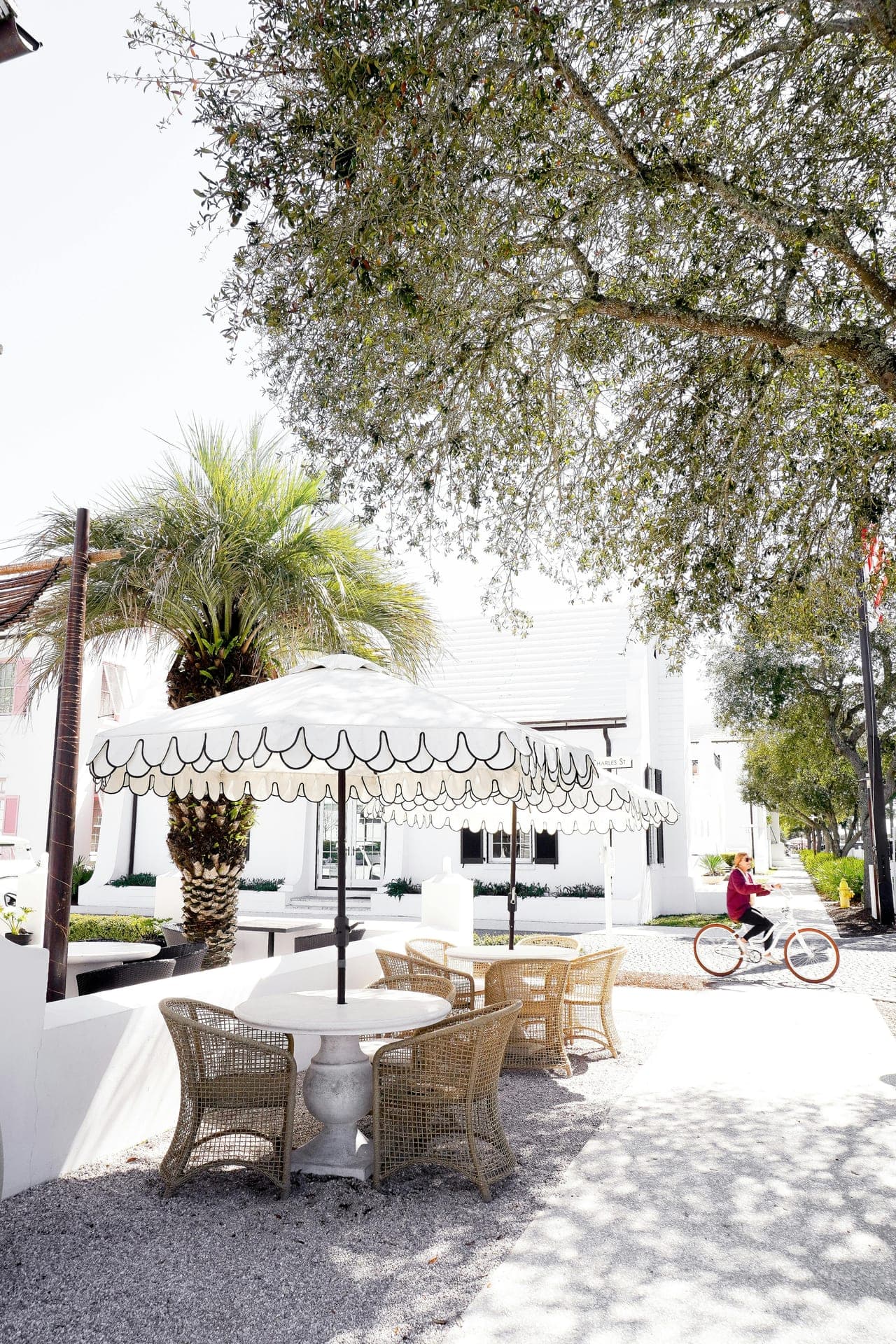 A scalloped white café umbrella in Seaside, Florida — palms, a person on a bicycle, the kind of vacation rental neighborhood we walk.