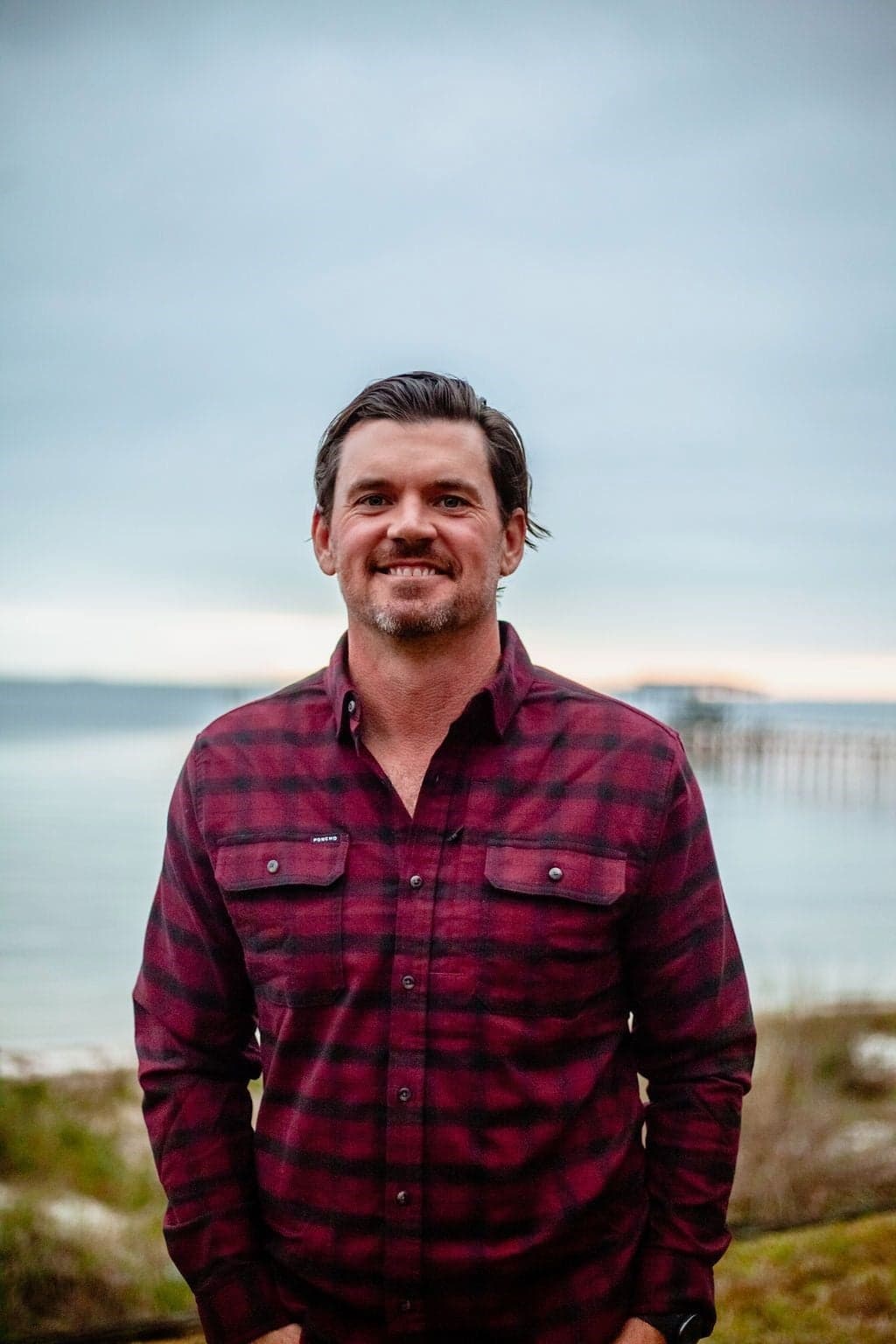 Harry Gibson, founder of VacayVerify, on the Emerald Coast shoreline at dusk — the Gulf and a pier behind him.