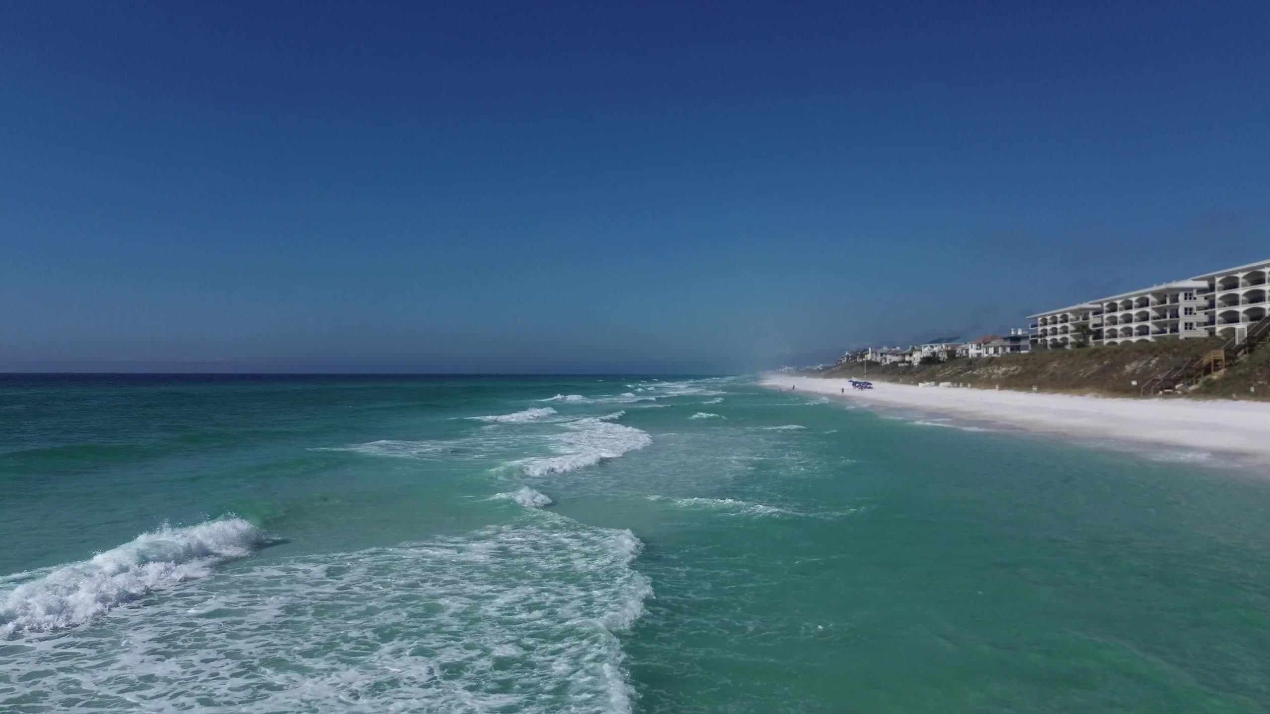An aerial view of the Emerald Coast — deep blue sky over vibrant emerald-green Gulf water rolling onto sugar-white sand.